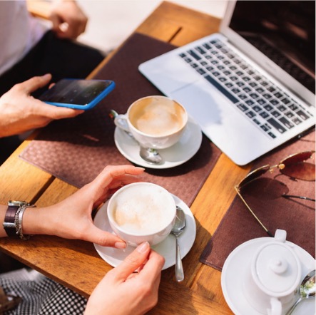 Hands holding cappuccinos at a cafe table with a laptop and phone